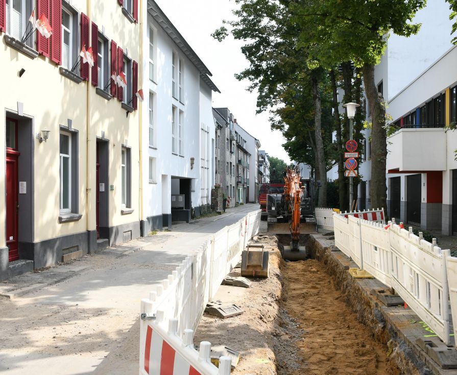 Straße mit Baustellenabsperrung. Bagger hebt Graben aus.